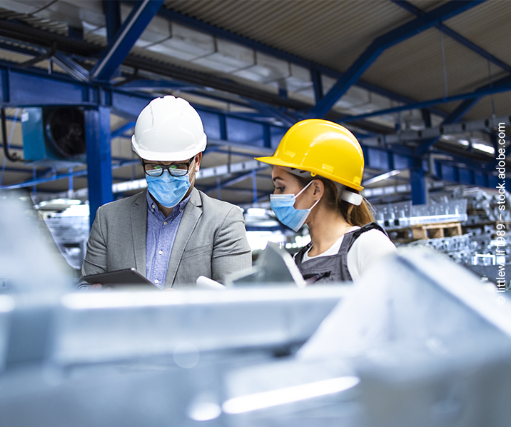 Zwei Personen mit Schutzhelm und Mund-Nasen-Maske stehen bei einer Besprechung in einer Industriehalle.