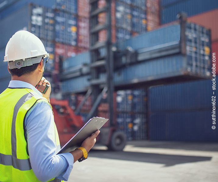 Man mit Bauhelm und Warnweste steht vor einer Containerwand. Er hält ein Funkgerät und ein Klemmbrett im Arm. 