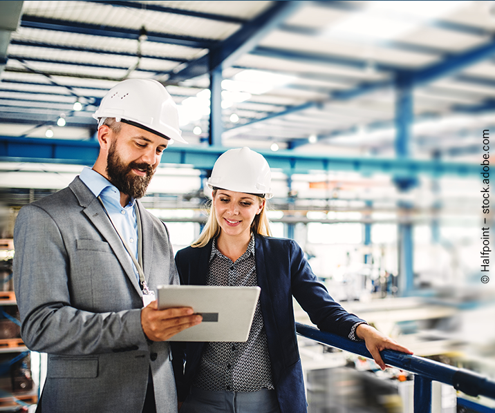 Mann und Frau mit Bauhelmen stehen in einer Fabrik und schauen auf ein IPad.