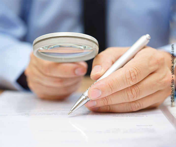 Eine Hand hält eine Lupe über einem Blatt Papier, die andere einen Stift. Webinar der IHK Südthüringen zum Prüfverfahren von Corona-Kurzarbeitergeldbezug