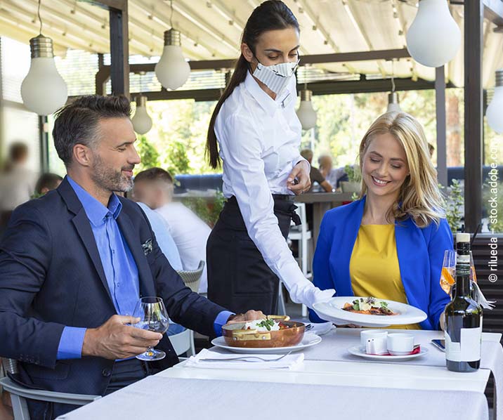 Ein Paar sitzt am Tisch im Restaurant und wird von einer Kellnerin mit Mund-Nasen-Schutz bedient.