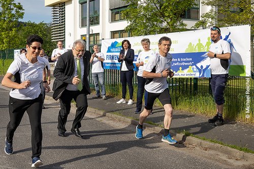 Läuferinnen und Läufer starten Lauf vor dem Hintergrund eines Firmengebäudes und klatschenden Personen Bild zum elften Thüringer Wald Firmenlauf