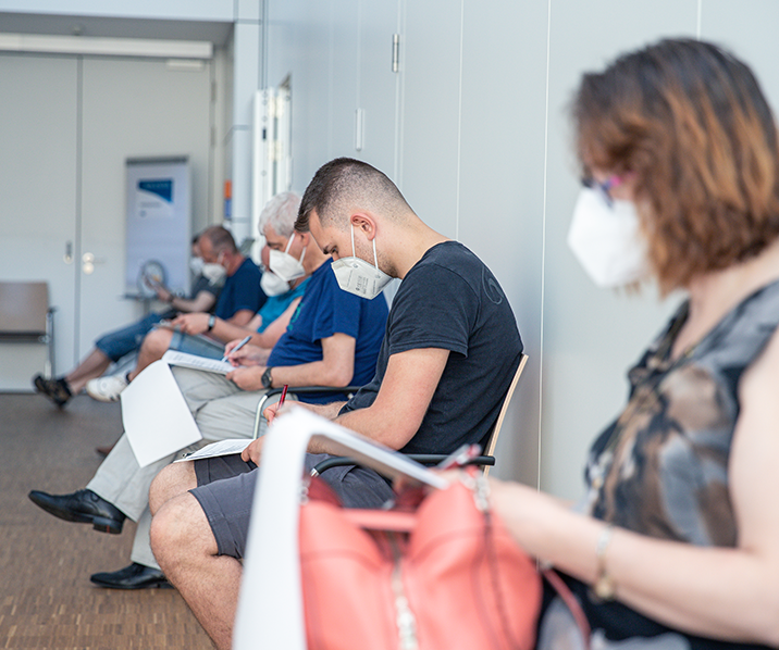 Menschen sitzen mit Mund-Nasen-Schutz in einer Reihe und füllen Fragebögen aus.